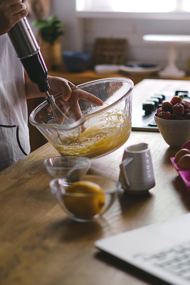 A woman uses an electric mixer to whip eggs in a cozy kitchen setting, surrounded by fruits and a laptop.
